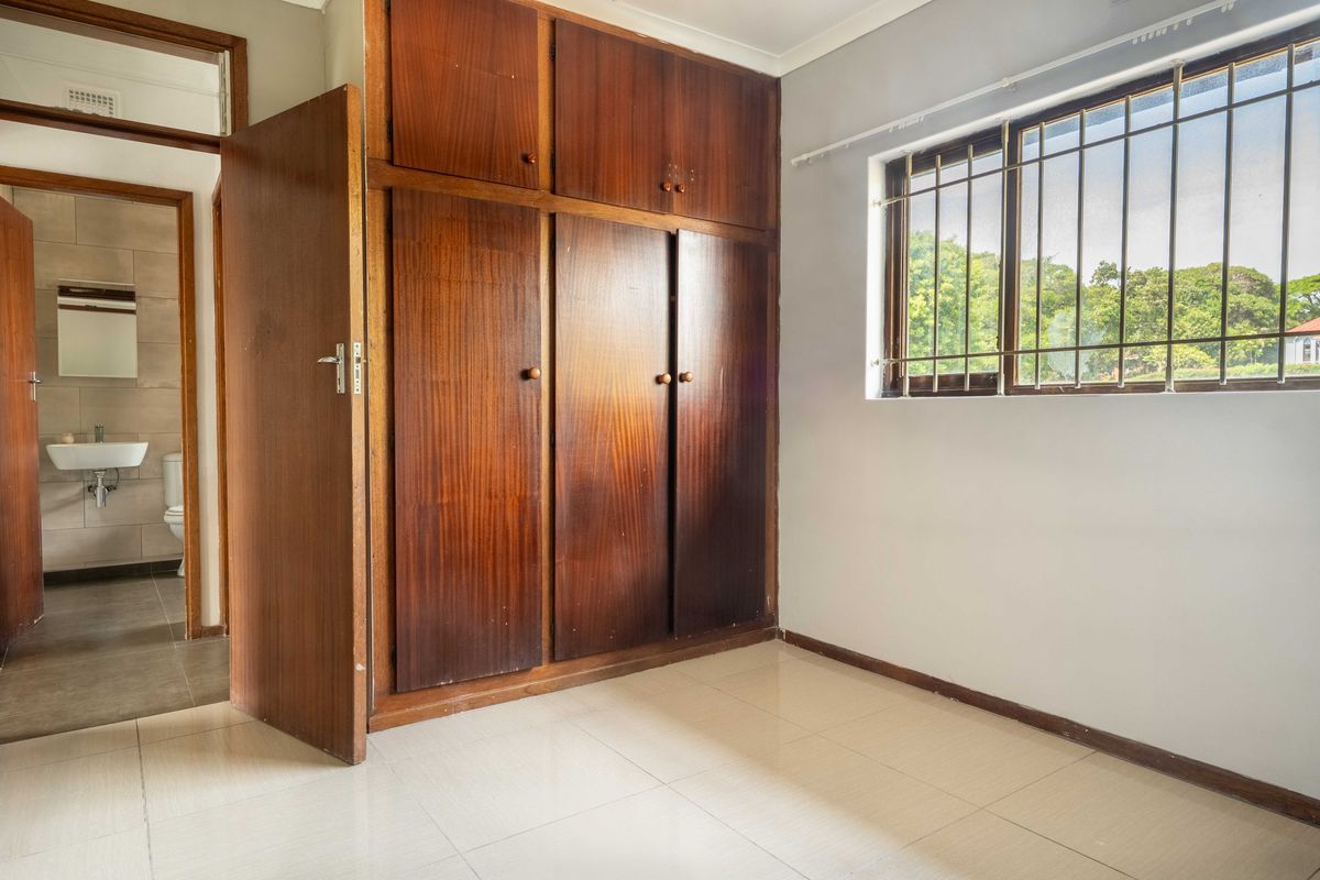 A bright, empty bedroom featuring built-in wooden cupboards, tiled flooring, and a window with security bars, with an adjacent bathroom visible.