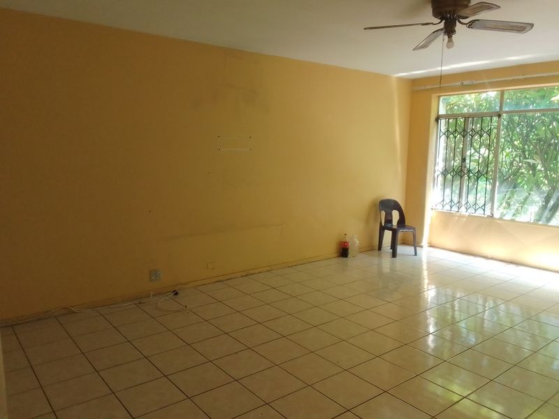 An empty living room featuring tiled flooring, a ceiling fan, and a large window with security bars, offering a blank canvas for renovation.