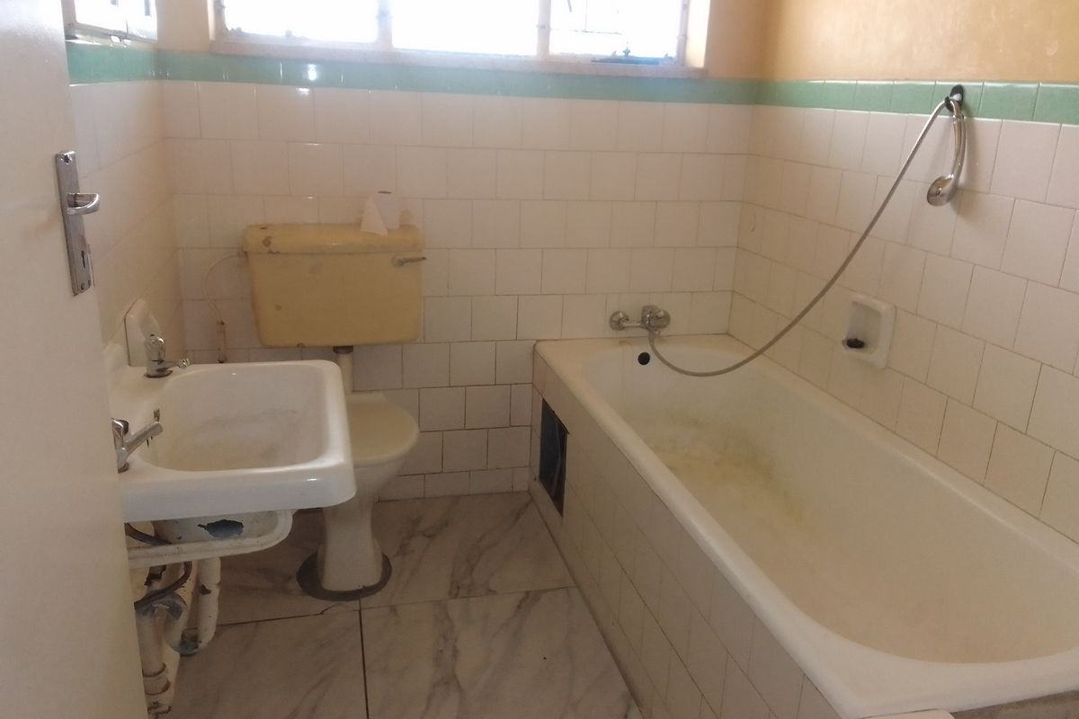 A dated bathroom featuring a pedestal basin, toilet with high-level cistern, and a bathtub with wall-mounted shower attachment, requiring modernization and cosmetic updates.