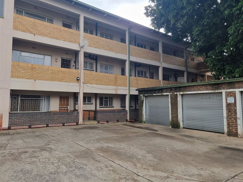 Exterior view of a multi-story apartment building featuring brick facade, balconies with security bars, and ground-level roller shutter garages with a concrete driveway.