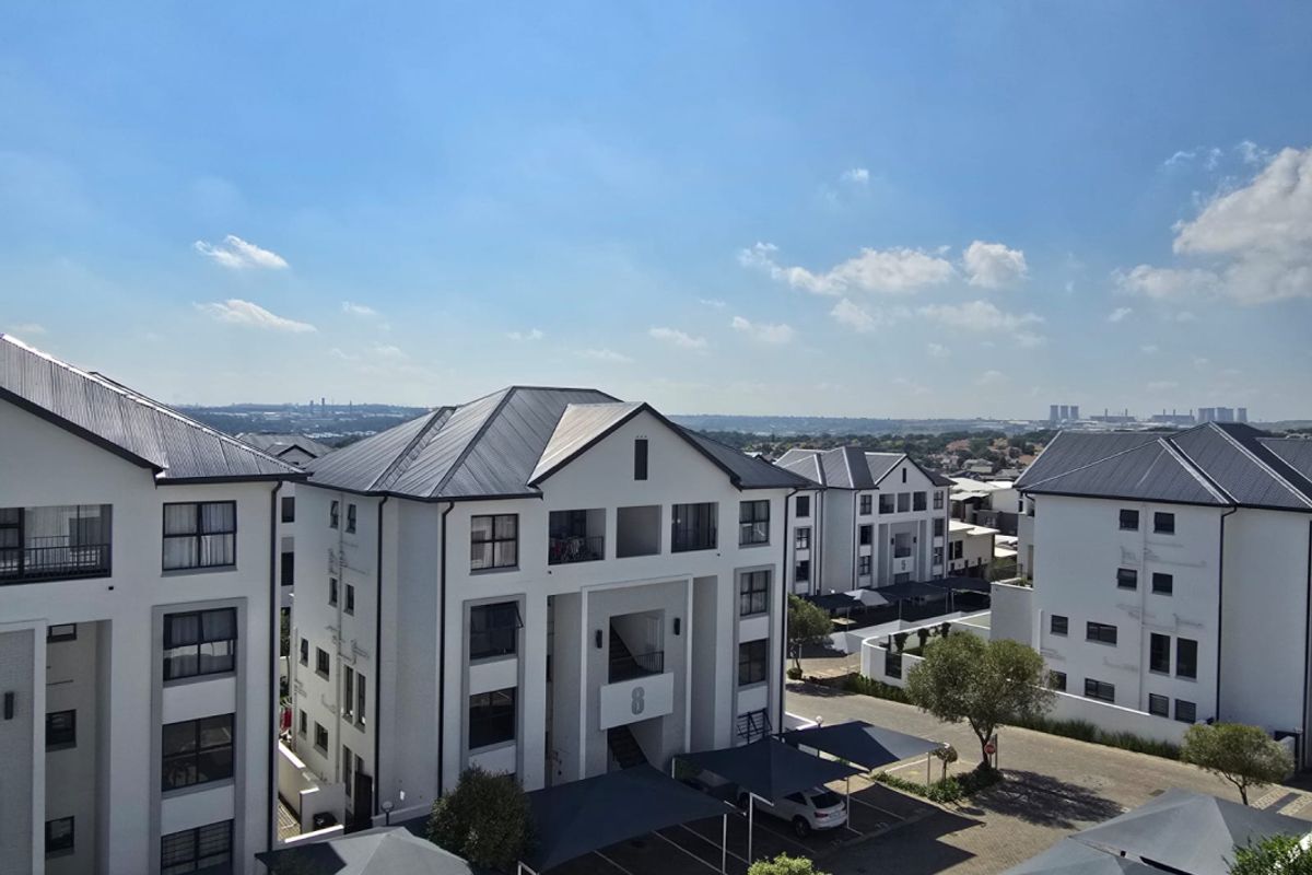 An elevated aerial view of a modern apartment complex featuring white facades, dark pitched roofs, and covered parking structures within a landscaped estate.