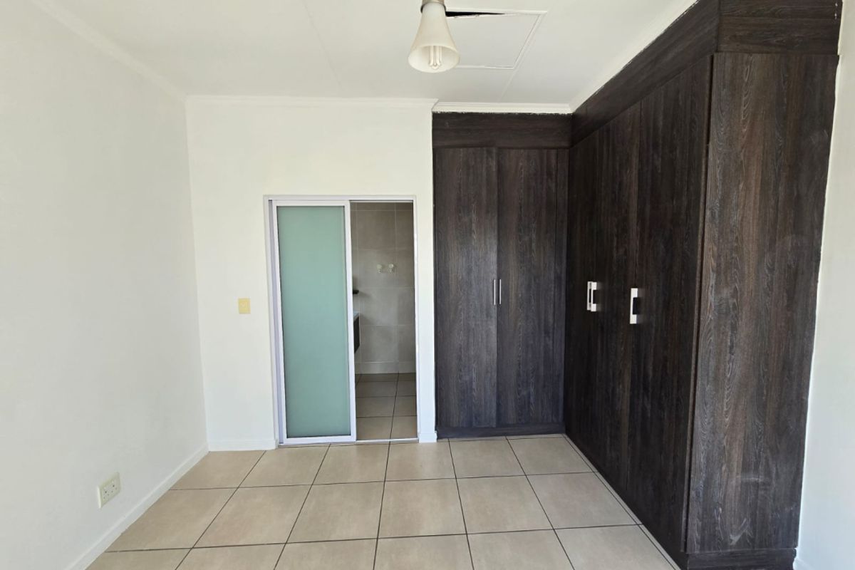 A clean, empty bedroom featuring dark wood built-in cupboards, neutral tiled flooring, and a sliding door leading to an en-suite bathroom.