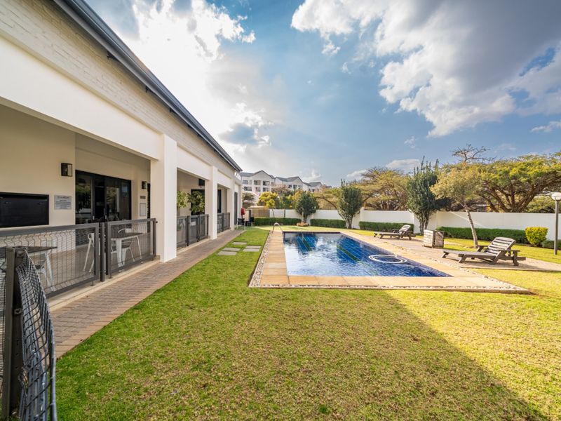 A well-maintained communal pool area featuring a paved deck, landscaped garden, and a covered patio with a built-in braai facility.