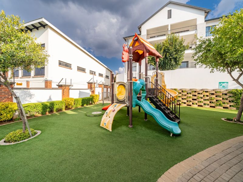 A well-maintained communal garden area featuring artificial turf, a children's playground structure, and secure perimeter walling with electric fencing.