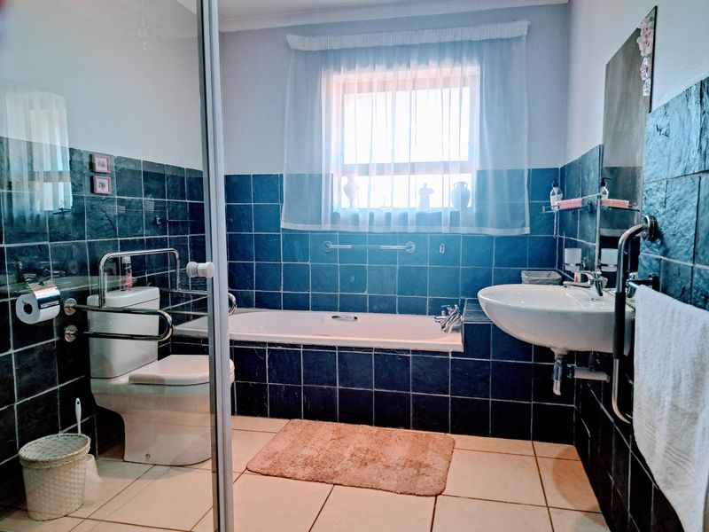A functional bathroom featuring dark tiled walls, a built-in bathtub, a wall-mounted basin, and a toilet equipped with safety grab rails.
