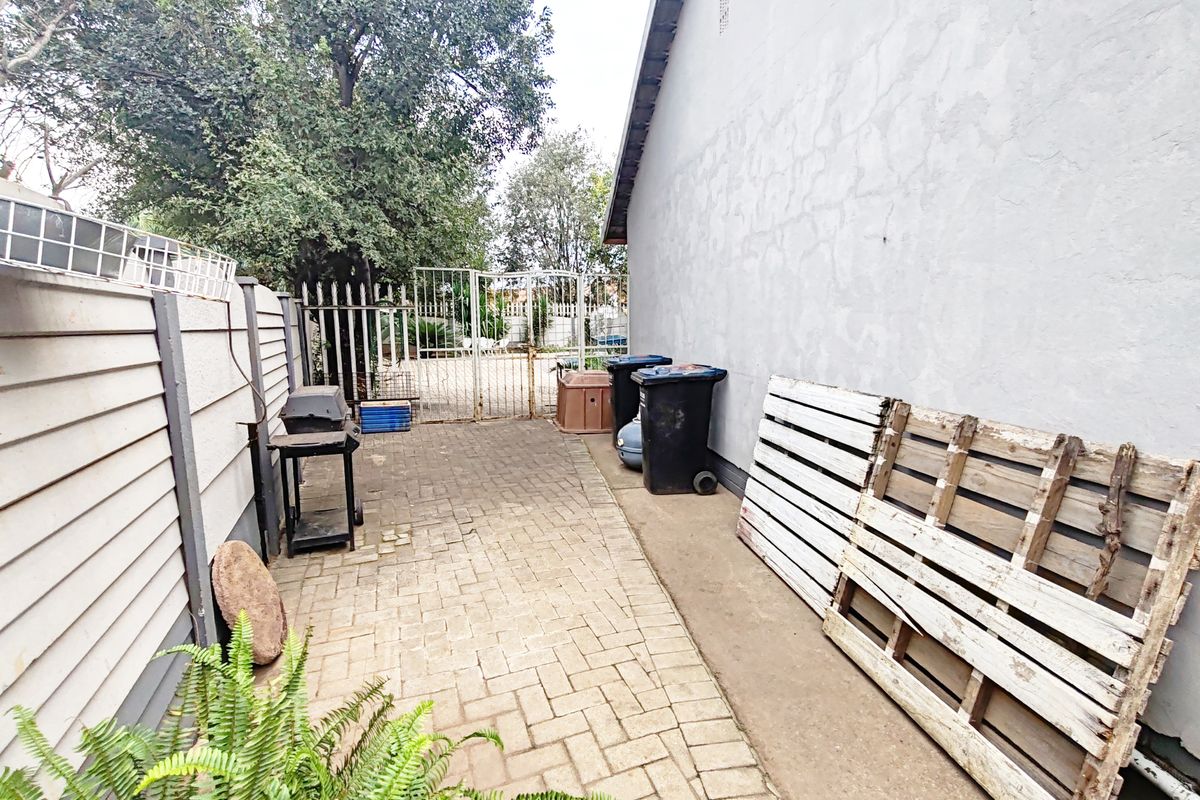 A side exterior walkway featuring paved flooring, perimeter walling, and a metal security gate, requiring maintenance to the wall plaster.