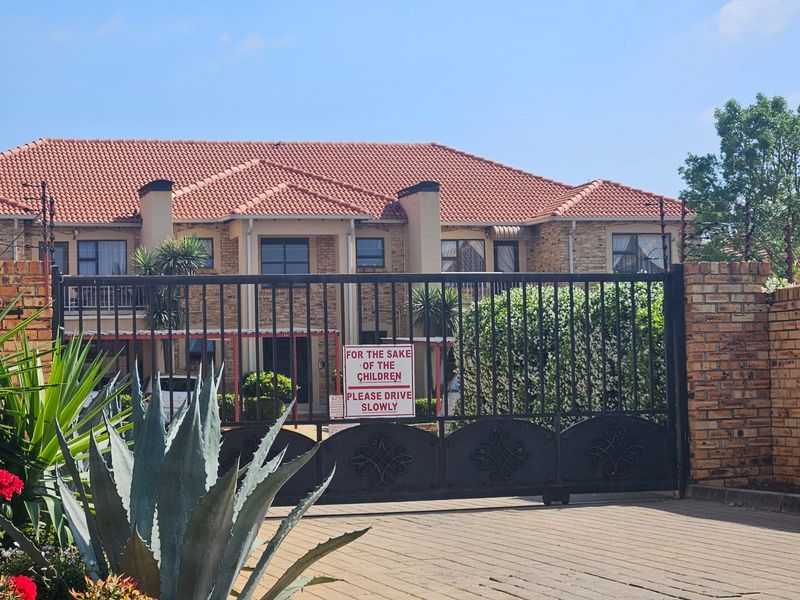 Front view of a multi-story apartment complex featuring a brick facade, tiled roof, and a secure gated entrance with paved driveway.