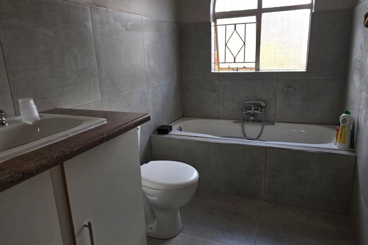 A functional bathroom featuring a built-in vanity, bathtub, and toilet with neutral grey wall and floor tiling and an arched window for natural light.