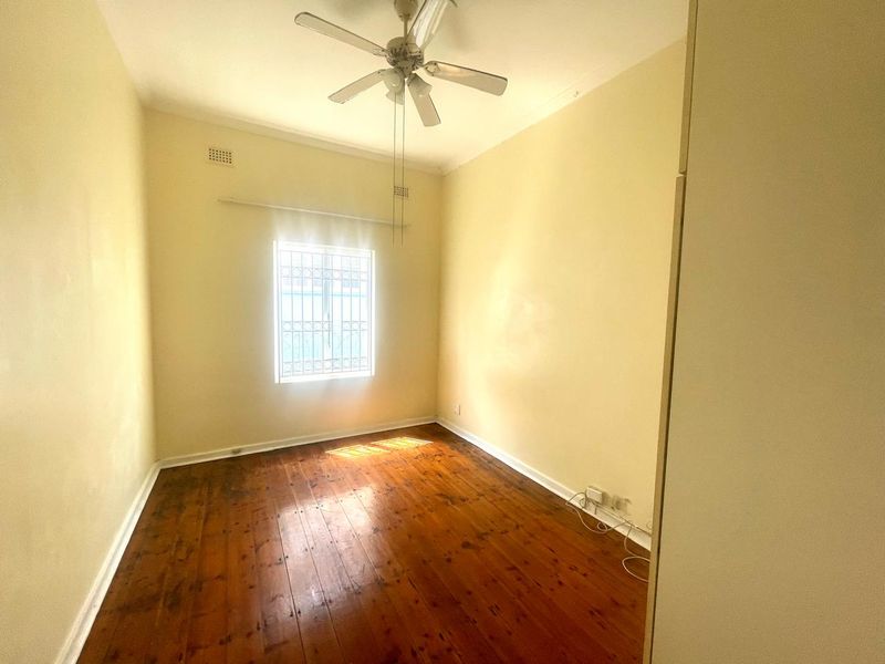 An empty bedroom featuring original hardwood flooring, a ceiling fan, and a window fitted with security bars.