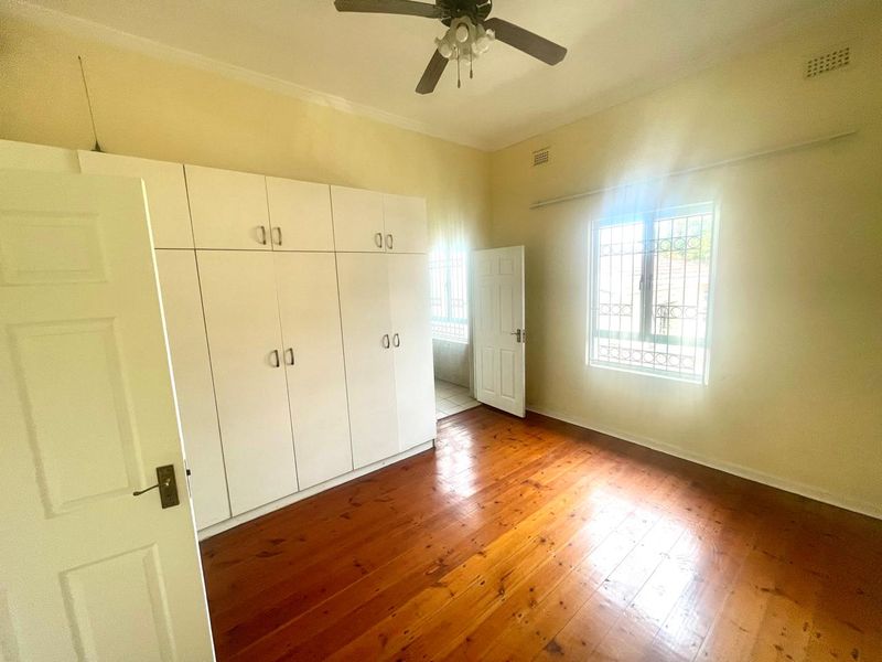 A bright, empty bedroom featuring polished timber flooring, built-in cupboards, and a ceiling fan, with security bars installed on the window.