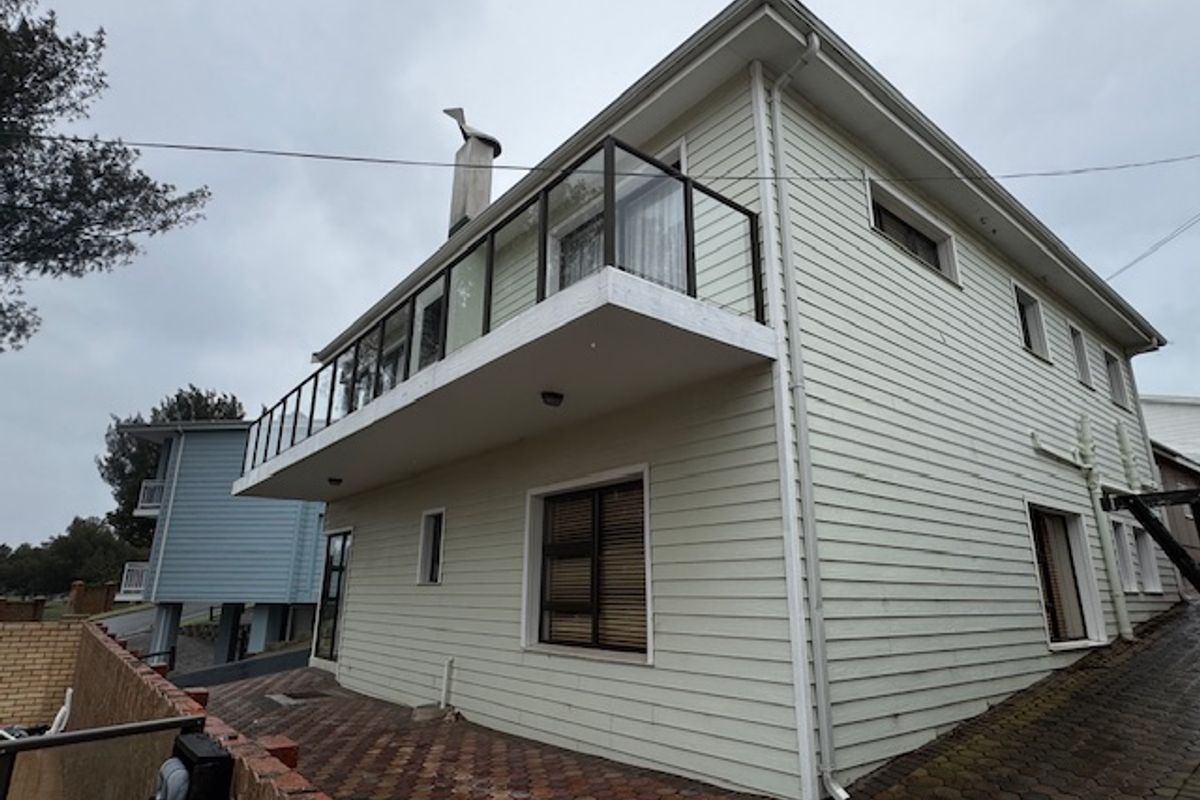 Rear exterior view of a two-story weatherboard house featuring a balcony with glass balustrade, chimney, and a paved patio area.
