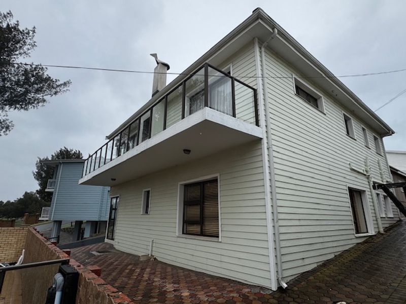 Rear exterior view of a two-story weatherboard house featuring a balcony with glass balustrade, chimney, and a paved patio area.