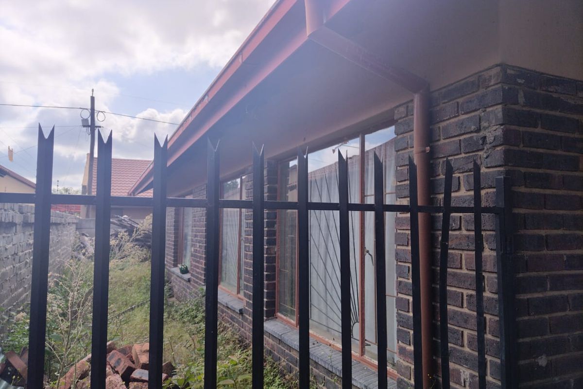 Rear exterior view of the property featuring face brick walls, metal security fencing, and window security bars, with overgrown garden area.