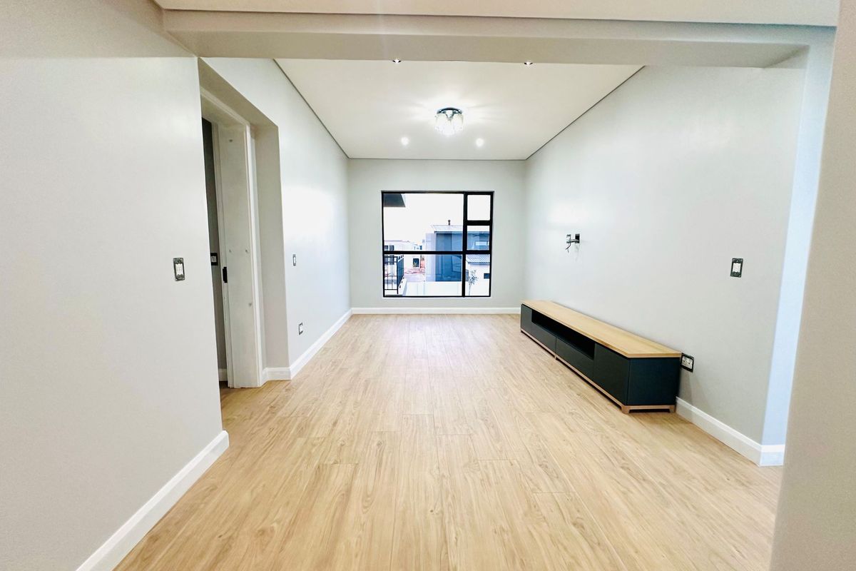 A bright, modern living room featuring light wood-look flooring, neutral walls, recessed ceiling lighting, and a large window providing natural light.