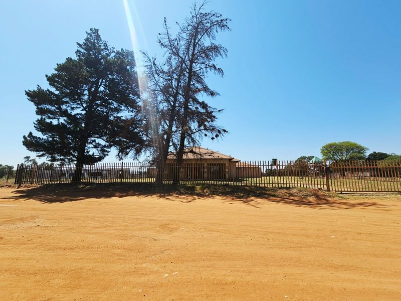 Street view of the property showing the dirt road access, perimeter metal fencing, and the main dwelling structure in the background.