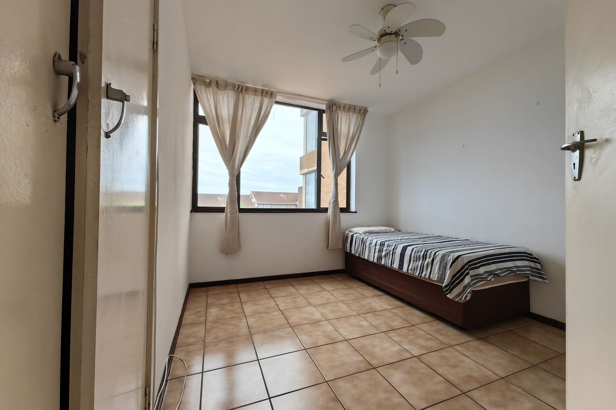 A simple bedroom featuring tiled flooring, a ceiling fan, and a large window providing natural light.
