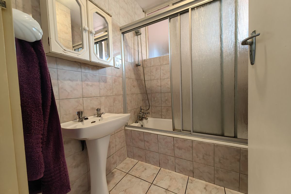 A dated bathroom featuring a pedestal basin, bathtub with a sliding shower screen, and full-height wall tiling.