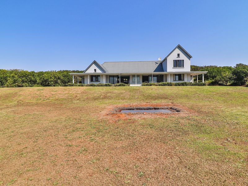 A rear view of a two-story farmhouse-style residence featuring a wrap-around veranda, metal roofing, and expansive surrounding grounds.