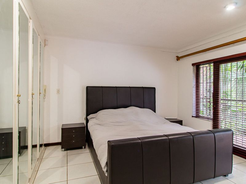 A bedroom featuring tiled flooring, a large built-in mirrored wardrobe, and a window fitted with security bars and venetian blinds.