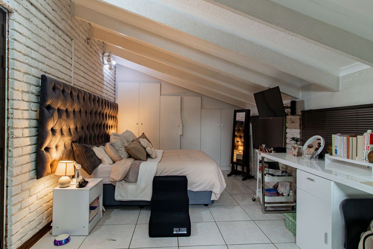A spacious bedroom featuring exposed ceiling beams, white painted brick walls, built-in storage cupboards, and tiled flooring.