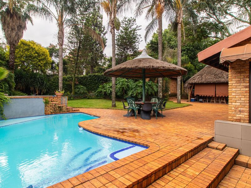 Outdoor entertainment area featuring a swimming pool, brick paved patio, and a thatched lapa set within a landscaped garden.