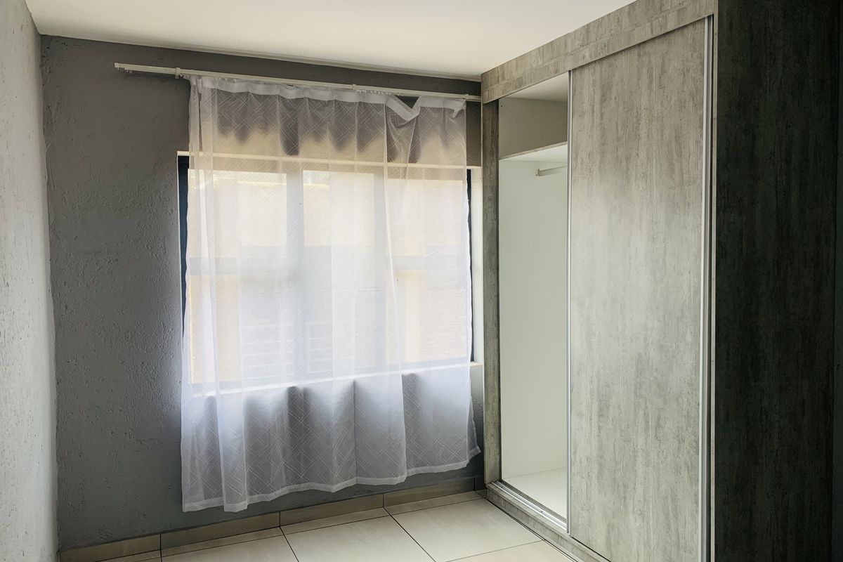 A bright, empty bedroom featuring modern grey textured built-in wardrobes, neutral tiled flooring, and a window with sheer curtains.