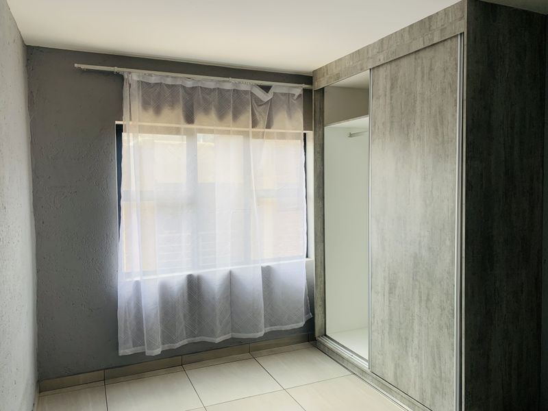 A bright, empty bedroom featuring modern grey textured built-in wardrobes, neutral tiled flooring, and a window with sheer curtains.