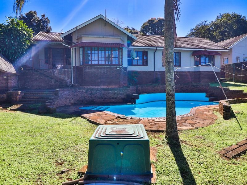 Rear view of the property featuring a swimming pool, brick retaining walls, a thatched lapa, and a spacious garden area.