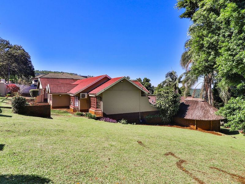 A view of the rear elevation of the house featuring a tiled roof, a separate thatched structure, and a spacious lawn area.