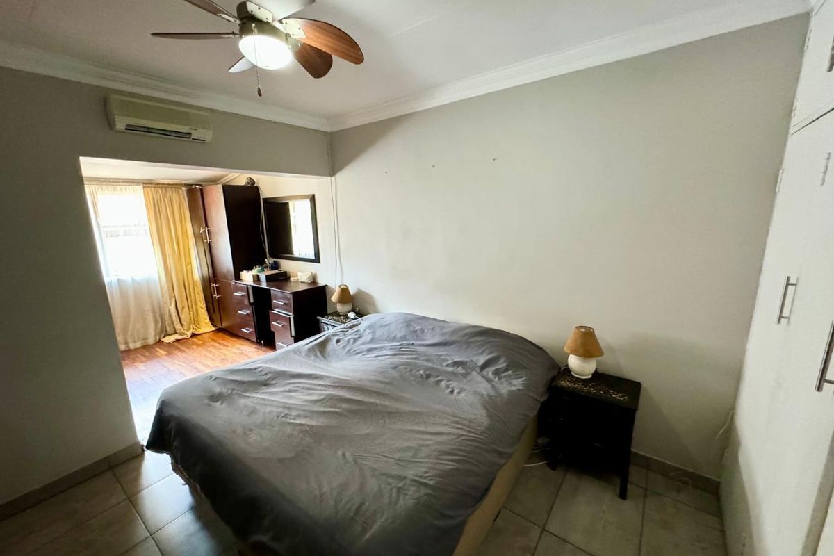 A bedroom featuring tiled flooring, a ceiling fan, a wall-mounted air conditioning unit, and built-in cupboards, with an adjoining alcove area.