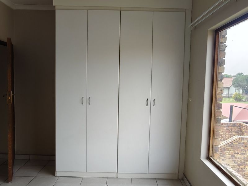 A bright, empty bedroom featuring built-in cupboards, tiled flooring, and a large window overlooking the exterior grounds.