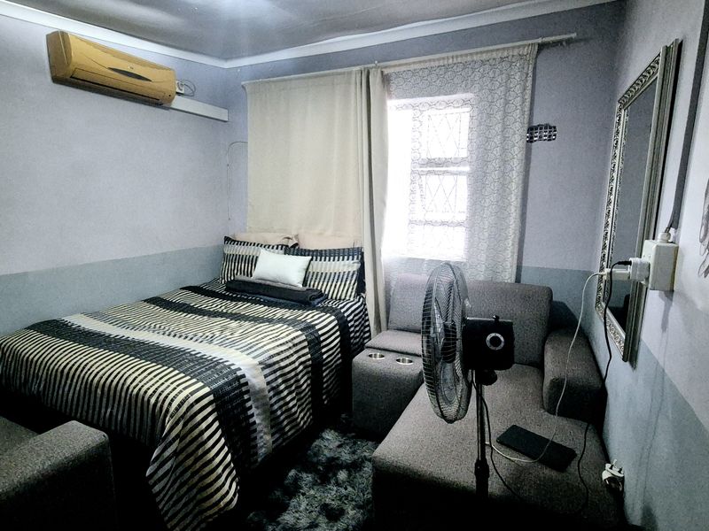 A compact bedroom featuring a wall-mounted air conditioning unit and security burglar bars on the window.