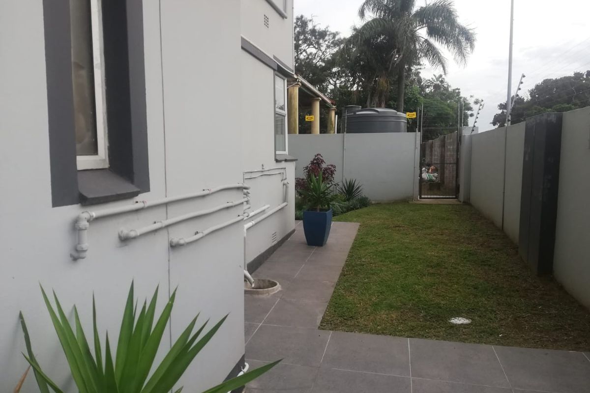 A side exterior view of the apartment featuring a paved walkway, a small lawn area, and perimeter boundary walls with security fencing.