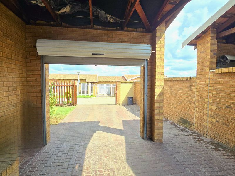 A covered garage area featuring exposed timber rafters, brick walls, and a roller shutter door, leading out to a paved driveway and boundary wall.