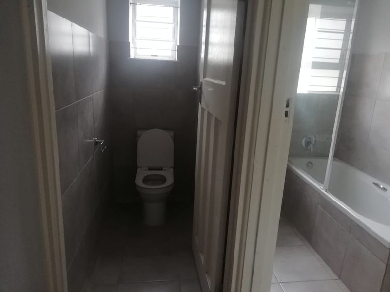 A functional bathroom featuring a separate toilet area and a bathtub with a shower screen, finished with neutral wall and floor tiles.
