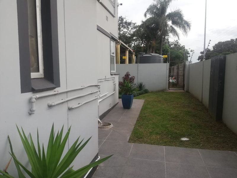 A side view of the apartment showing a paved patio area, a small lawn, perimeter wall with electric fencing, and an external water tank.