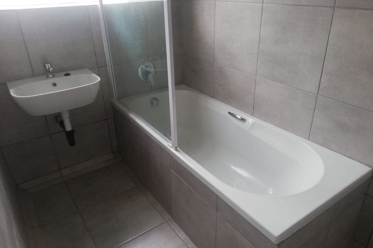 A clean, modern bathroom featuring a built-in bathtub with a glass shower screen and a wall-mounted basin with neutral ceramic tiling throughout.