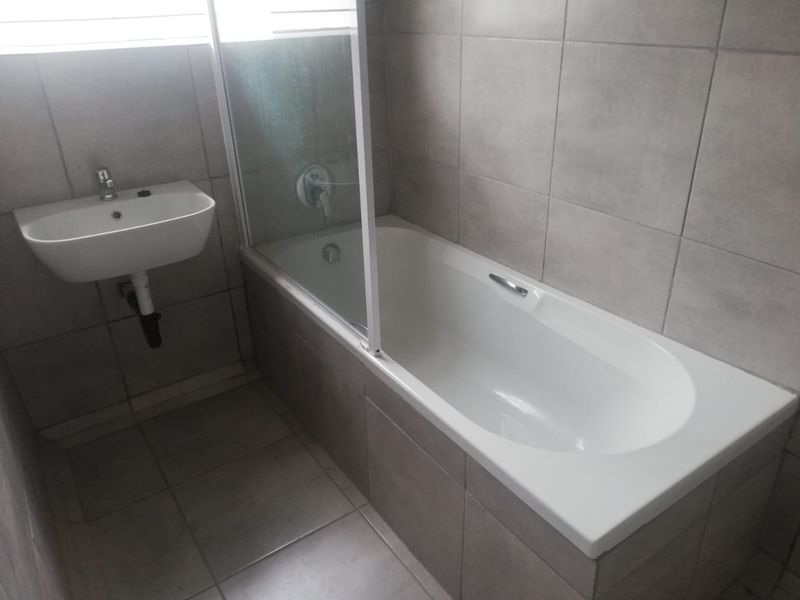 A clean, modern bathroom featuring a built-in bathtub with a glass shower screen and a wall-mounted basin with neutral ceramic tiling throughout.