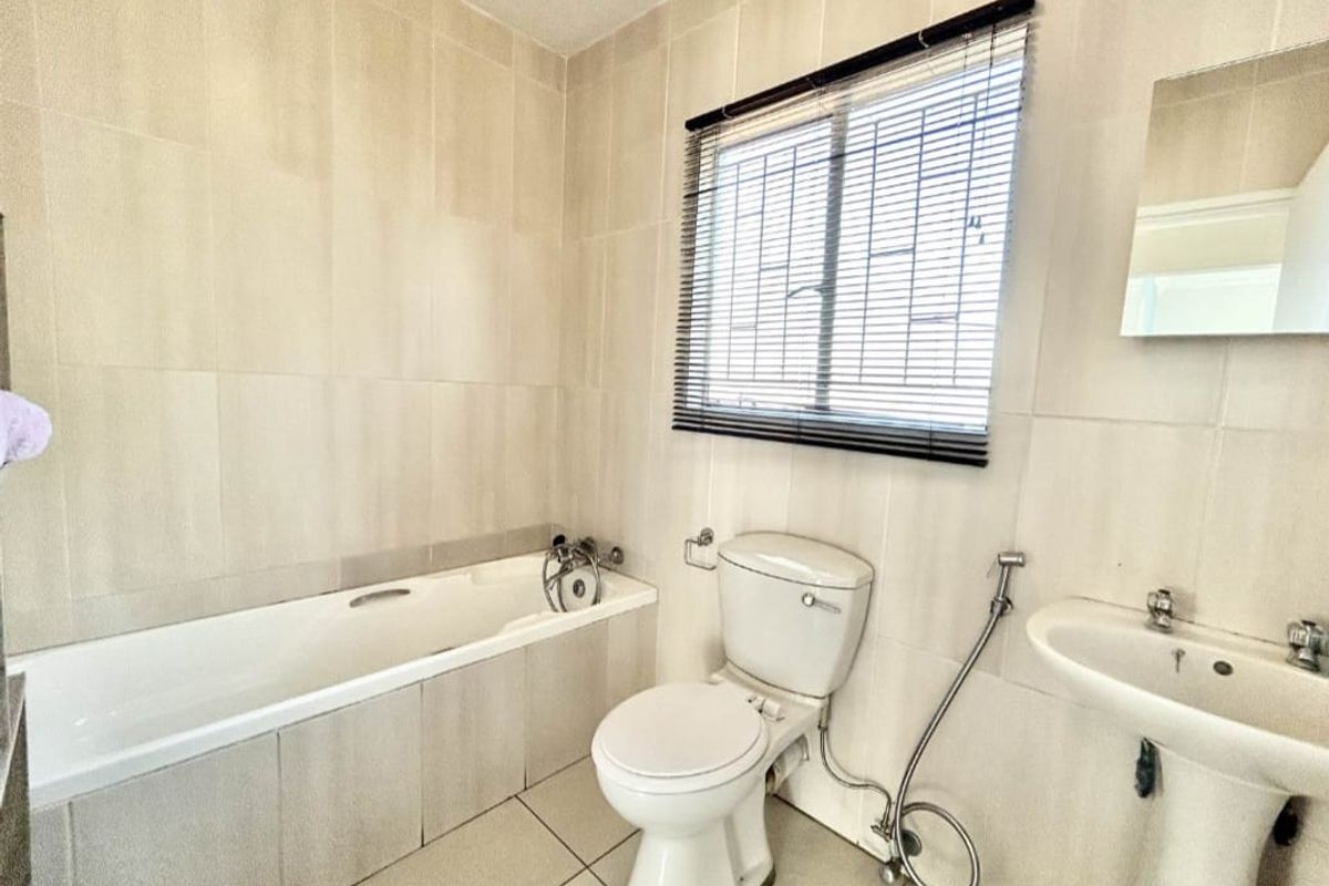 A functional bathroom featuring a built-in bathtub, pedestal basin, and toilet, finished with neutral wall and floor tiling and a security-grilled window.