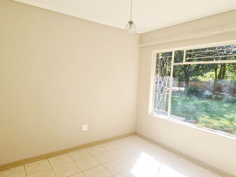 A bright, empty bedroom featuring tiled flooring, built-in cupboards, and large windows with security bars overlooking the garden.