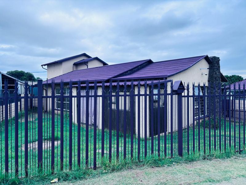 Front view of a residential house featuring a metal security fence, stone chimney, and a corrugated metal roof.
