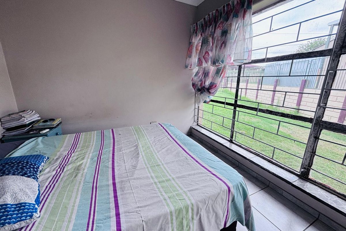 A bedroom featuring tiled flooring and a large window fitted with security bars, offering a view of the exterior garden area.