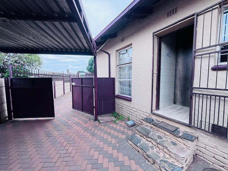 Rear exterior view featuring a covered carport, paved driveway, security gates, and stone steps leading to a tiled entrance area.