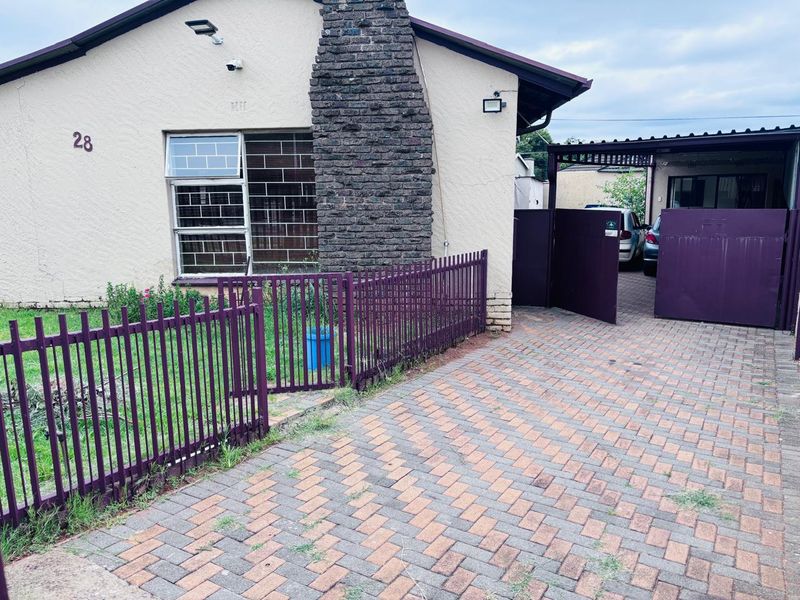 Front elevation of a house featuring a brick chimney, paved driveway, carport, and secure metal fencing with security bars on the windows.