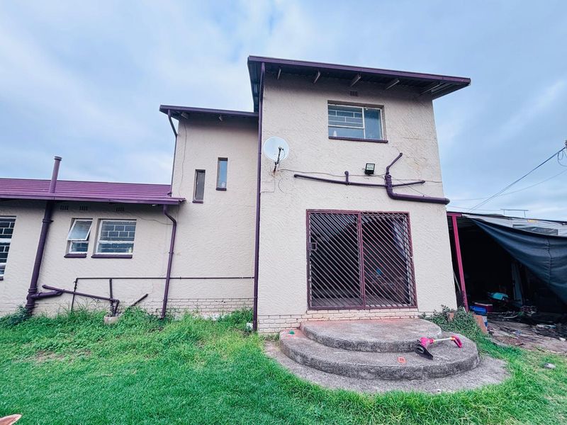 Rear view of a two-story house featuring a concrete entrance, metal security gate, and external plumbing fixtures set against a large garden area.