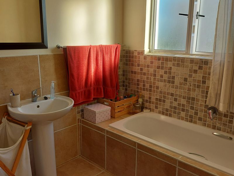 A functional bathroom featuring a pedestal basin, a built-in bathtub with tiled surround, and neutral floor tiling, illuminated by natural light from a frosted window.