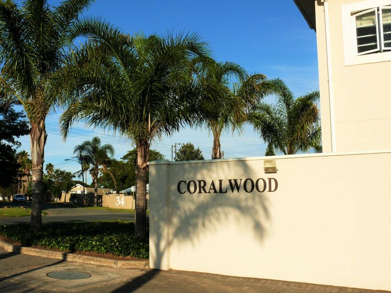 The entrance to Coralwood complex featuring a prominent boundary wall, mature palm trees, and paved driveway access.
