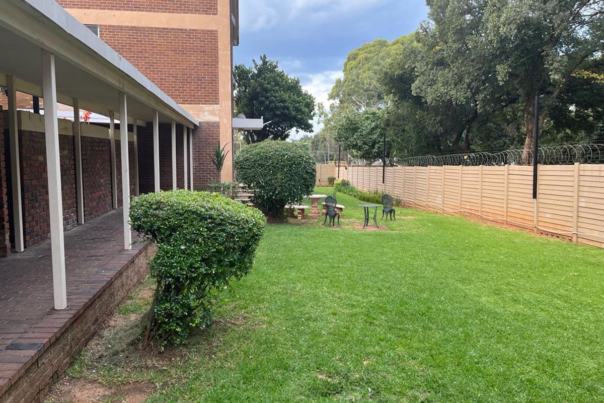 A rear exterior view of the flat featuring a covered brick walkway, a well-maintained lawn, and a secure perimeter fence with razor wire.