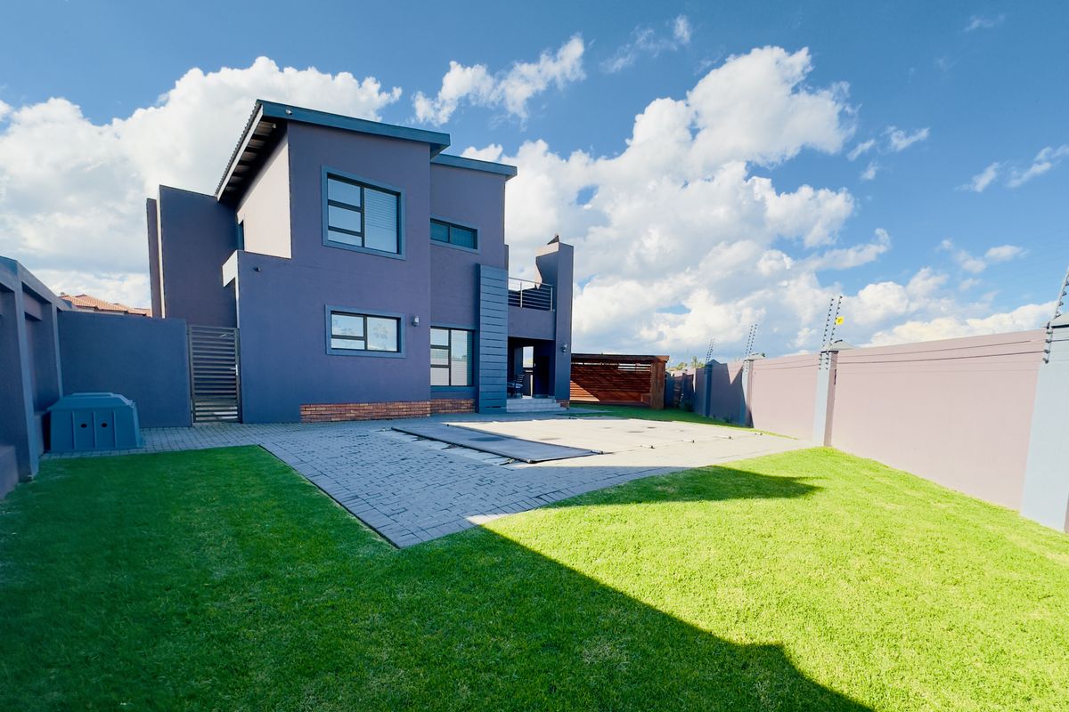 Rear view of a modern double-story home featuring a paved patio, swimming pool with cover, landscaped lawn, and secure perimeter wall with electric fencing.