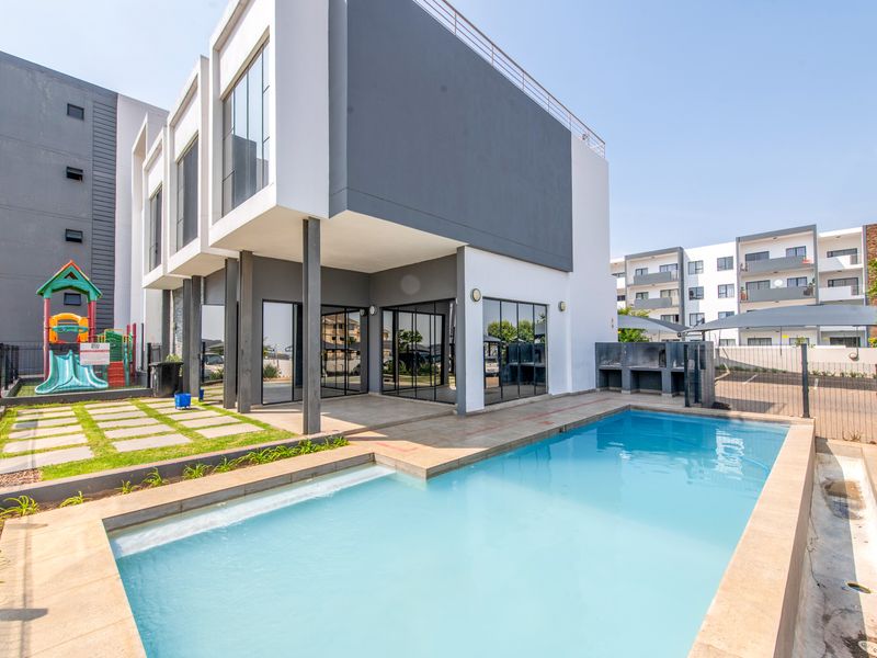 Modern apartment complex exterior featuring a swimming pool, paved patio area, and built-in braai facility, set within a contemporary residential development.